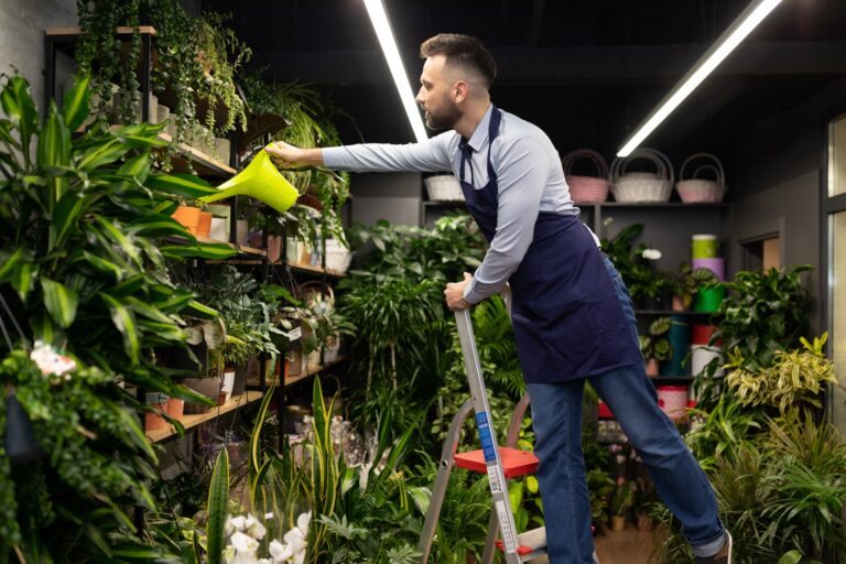 a male gardener in a flower shop standing on a ladder watering flowers on the shelves